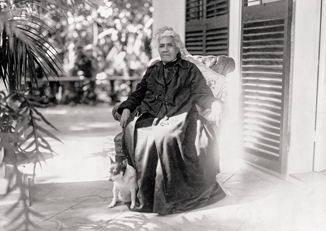 black-and-white archival photo of robed woman sitting in chair on outdoor patio next to small dog, with louvered doors on right and plants on left