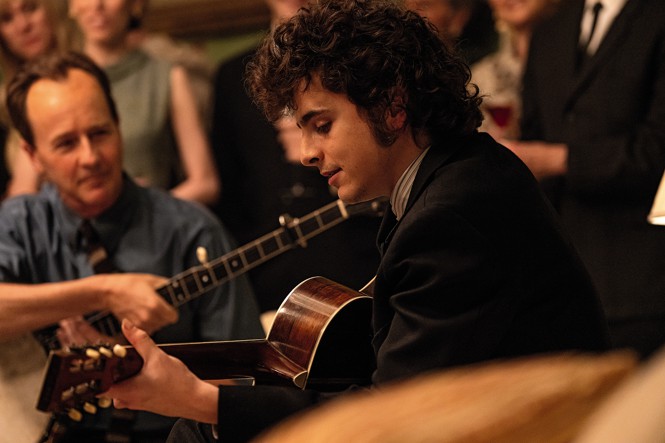 movie still with Chalamet in character as Bob Dylan strumming a guitar with crowd behind