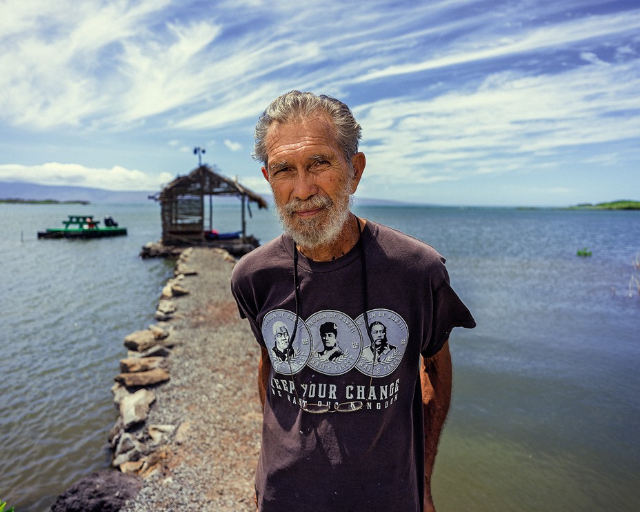 photo of bearded man wearing t-shirt showing Hawaiian coins and words "KEEP YOUR CHANGE WE WANT OUR KINGDOM" standing on stone pier surrounded by water and sky