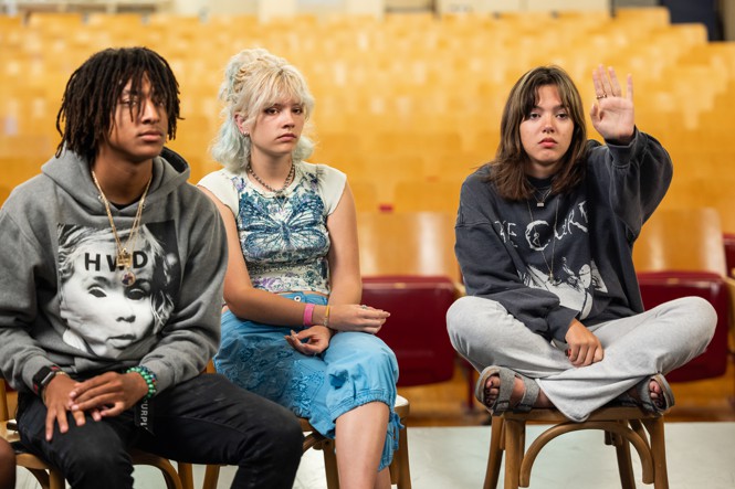 Three students sit in an auditorium and the girl on the right has her hand raised