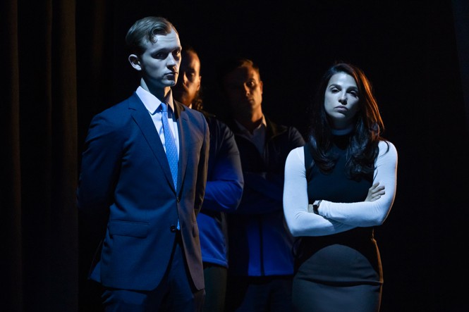 A man in a suit and a woman with a black dress and her arms crossed stand in a dark blue light