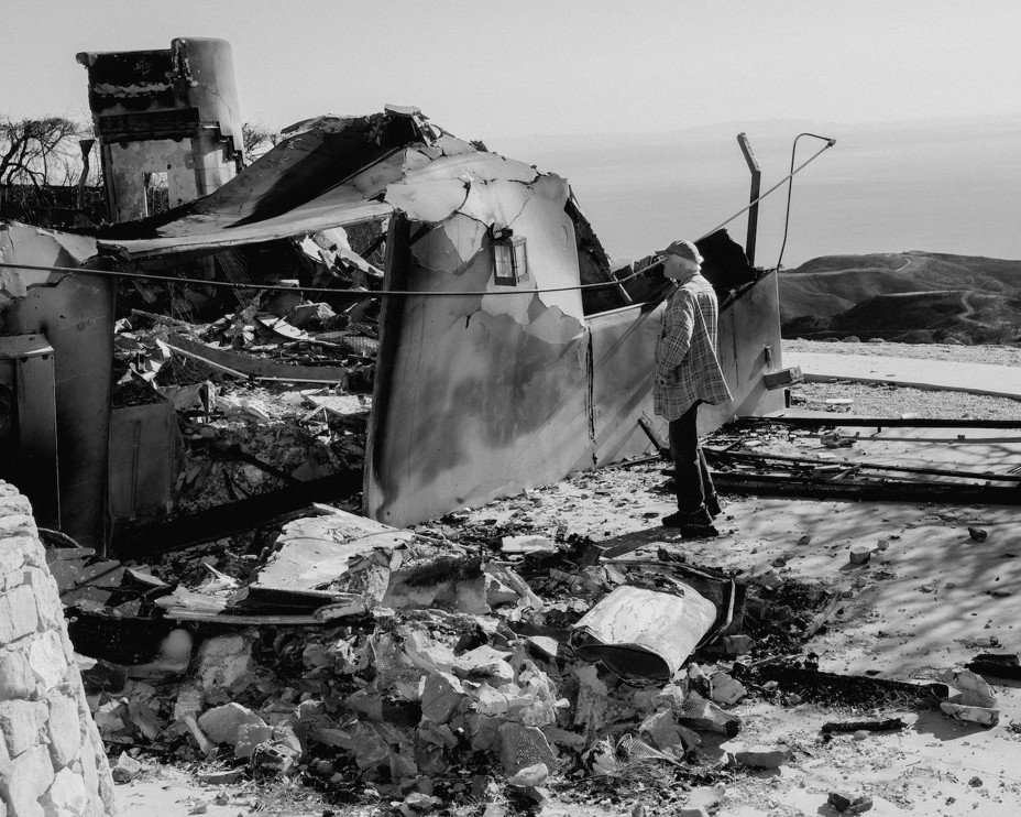 In a black and white photo a man looks at a burned down home