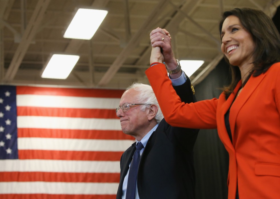 Bernie Sanders and Tulsi Gabbard hold arms up in the air with an American flag behind them