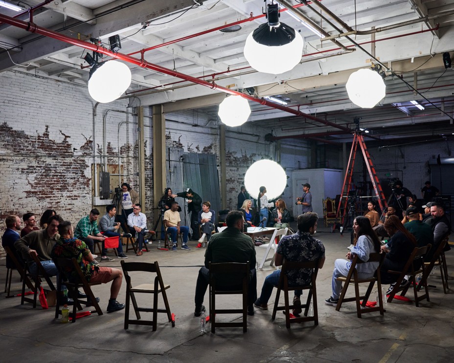 A group of people sit around in a circle in chairs while they are filmed 