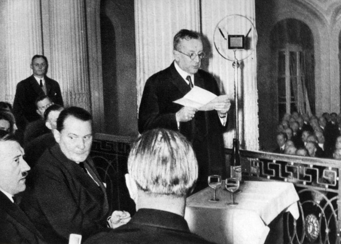 In a black and white photo Fritz Thyssen speaks at a microphone in front of Hitler and others