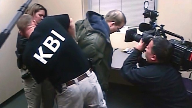 In a photo there is a man wearing a KBI shirt arresting another man while someone films 