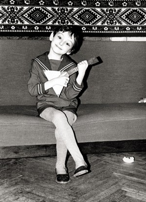 black-and-white photo of smiling child in sailor suit sitting with legs crossed and playing an instrument