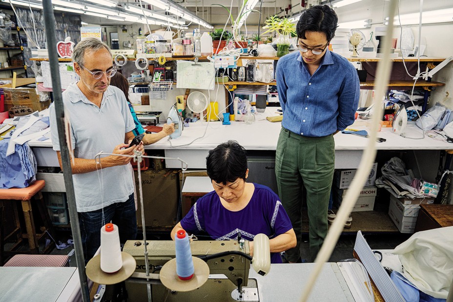 photo of author and another man standing behind a woman sitting at a sewing machine in a factory, watching her work