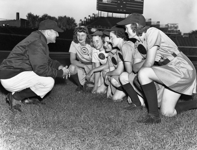 black-and-white photo of male coach squatting and talking to group of 5 women in skirted baseball uniforms kneeling on field and listening to him