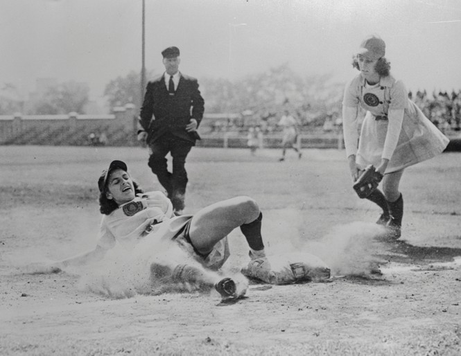 black-and-white photo of woman in baseball uniform sliding into base with opposing player attempting the tag and umpire in background