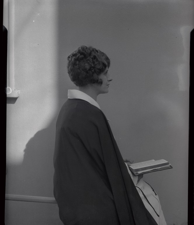 In a black and white photo a woman sits on a stool facing towards the right.