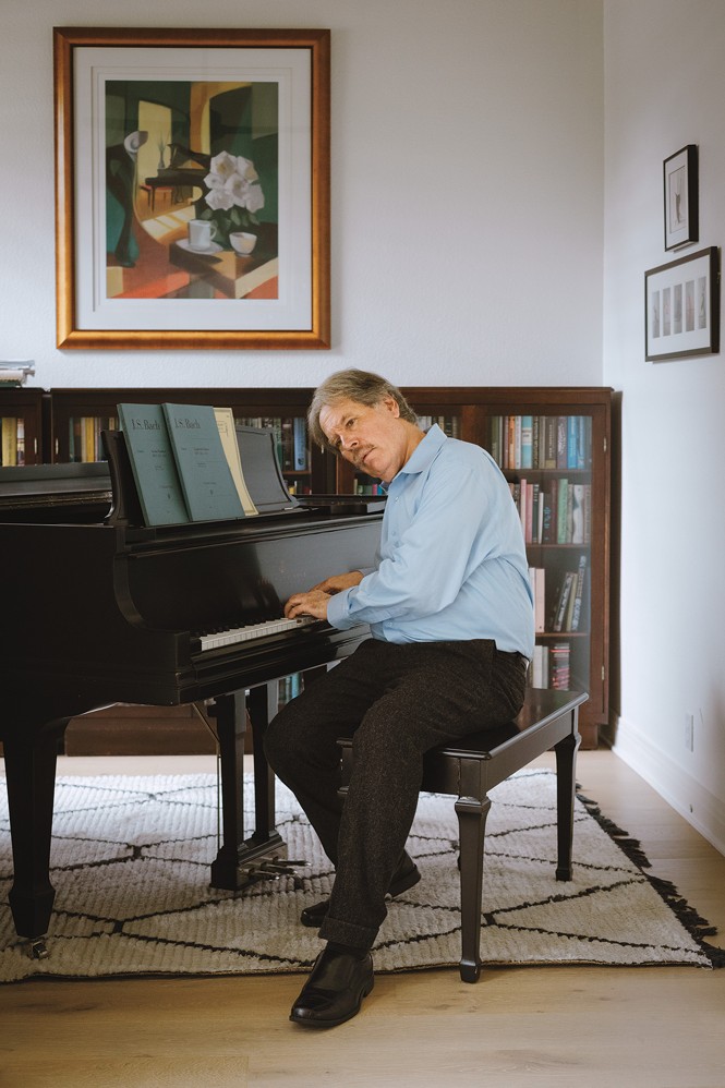 photo of man sitting at piano with hands on keyboard, books and artwork in background