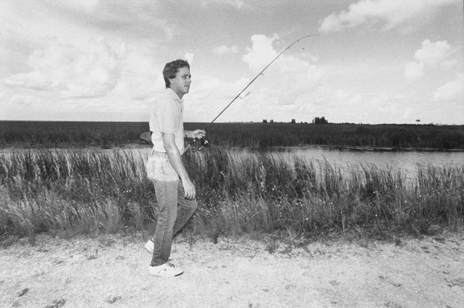 black-and-white photo of man in jeans wet to the upper thigh walking next to tall grasses along shoreline with fishing pole