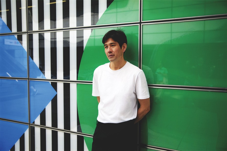 photo of man in white tee shirt and black pants standing against wall with graphic mural of blue square, green circle, and black-and-white stripes 