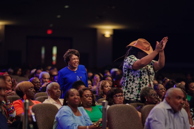 Two people in a crowd of seated people stand up smiling and clapping