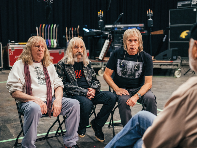 photo of 3 bandmates sitting in folding chairs and facing interviewer on stage with equipment and amps behind