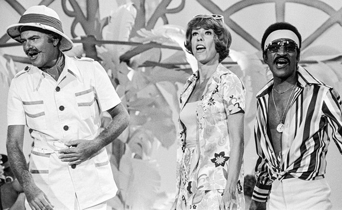 black-and-white photo of 3 people singing on stage: Korman in white safari outfit and fake mustache, Burnett in print dress, and Davis Jr. in headband, sunglasses, and open striped shirt