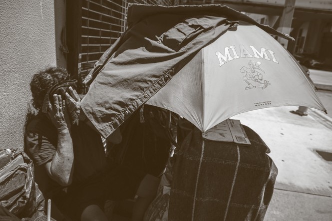 black-and-white photo of person sitting, holding a cloth to their face, leaning against a wall with suitcase and large umbrella with MIAMI on it