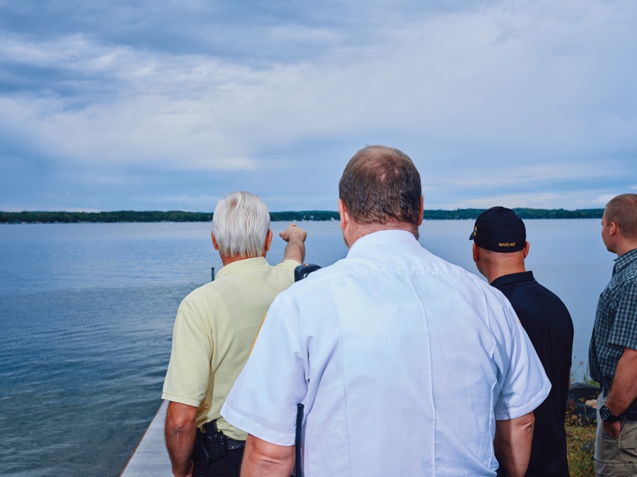 photo from behind of 4 men standing on dock at lake, one pointing toward the opposite shore in the distance