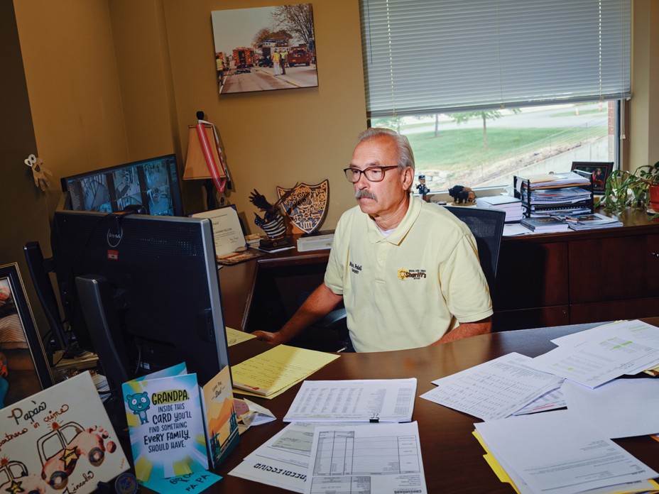 photo of man wearing glasses and polo shirt sitting at computer behind desk strewn with papers and cards, with window in background