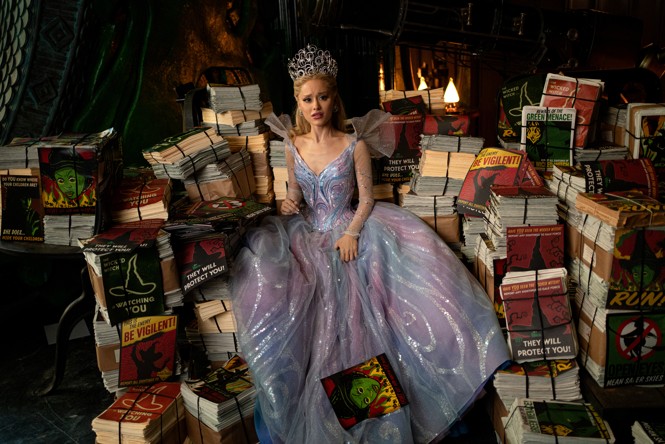 A color still from Wicked: For Good of Glinda sitting amongst a pile of books in a dark room
