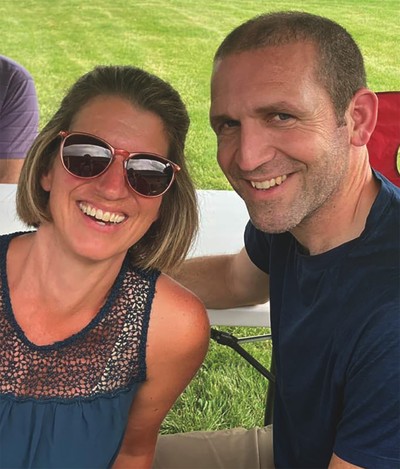 photo of woman in sunglasses and man with arm around her smiling with table and lawn in background
