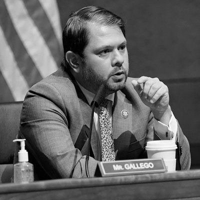 black-and-white photo of man speaking at mic while seated in Congress with American flag behind