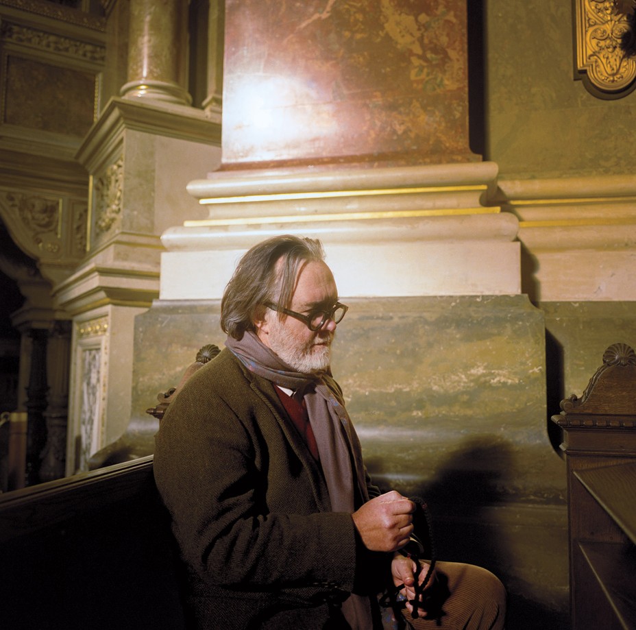 photo of beaded man in glasses holding prayer beads and sitting in a pew inside an ornate marble wall