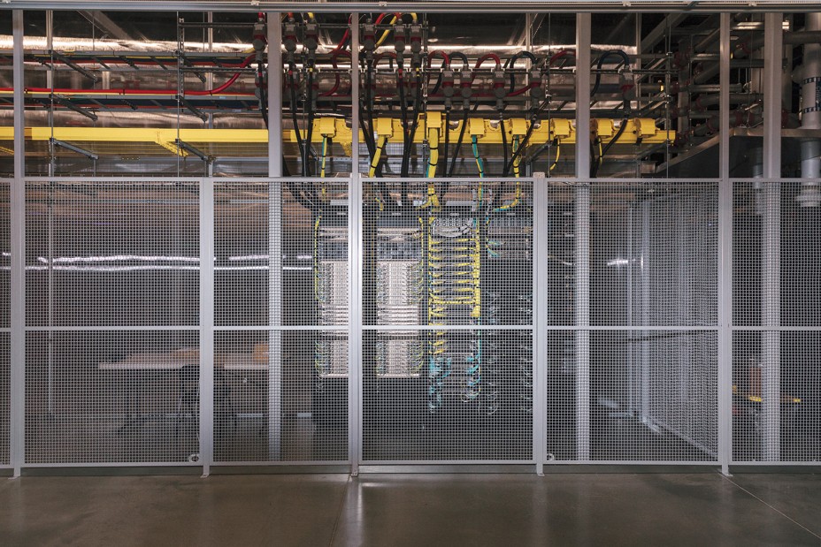 photo from inside warehouse of metal mesh cage around stacks of computer equipment with numerous cables extending to ceiling