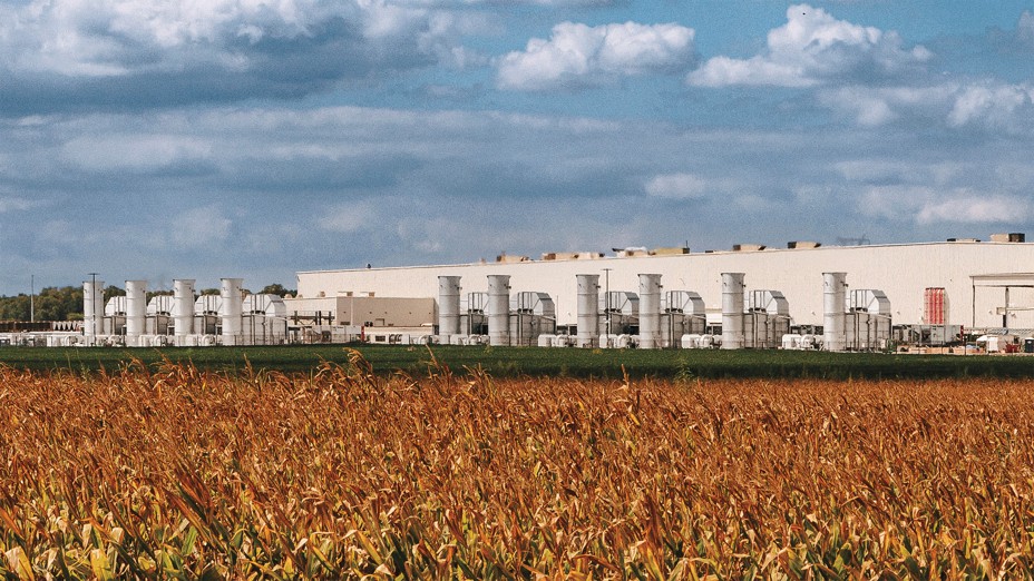 photo of enormous warehouse with numerous external cooling structures, with bronzed field of corn growing in foreground