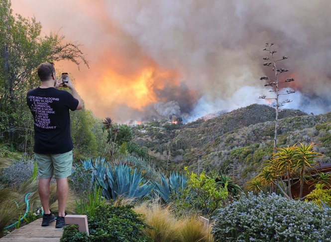 Photograph of Spencer Pratt recording a fire burning on hills nearby with his cellphone. He is wearing green shorts and a black t-shirt.