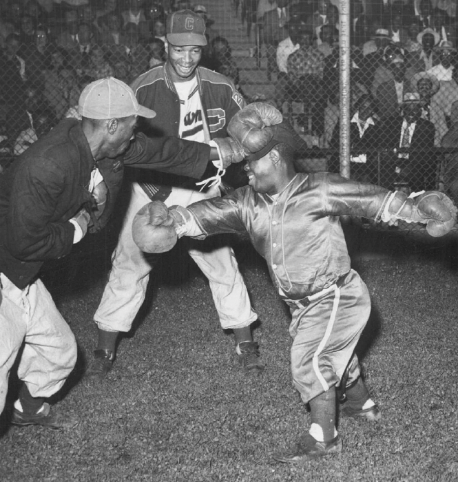 black-and-white archival photo of man in baseball uniform wearing boxing gloves holding the head of a little person in baseball uniform and boxing gloves at arm's length