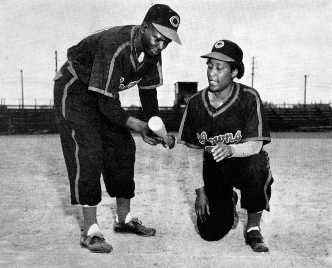 black-and-white archival photo of woman in baseball uniform on one knee talking to man in baseball uniform bent at the waist