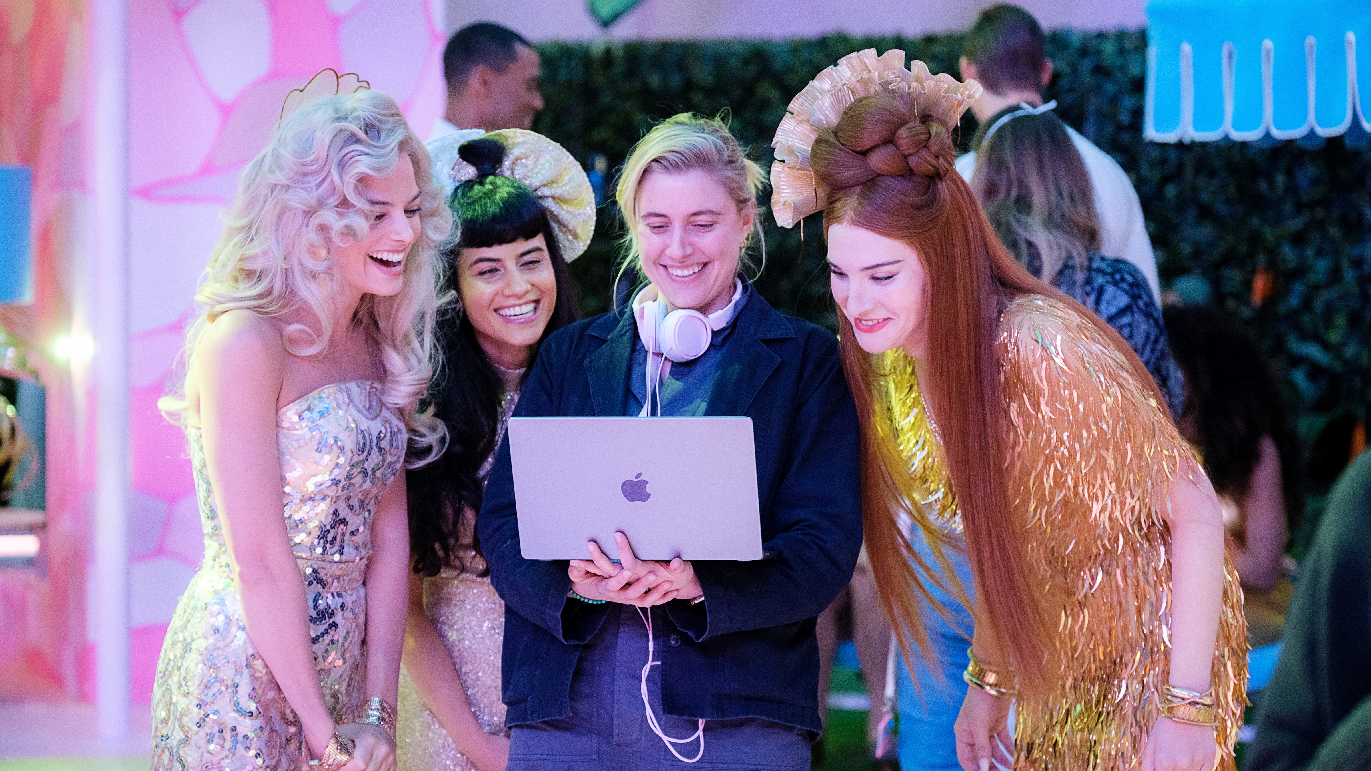 An image of Greta Gerwig on the set of Barbie, looking at a laptop, surrounded by Barbie stars (including Margot Robbie)