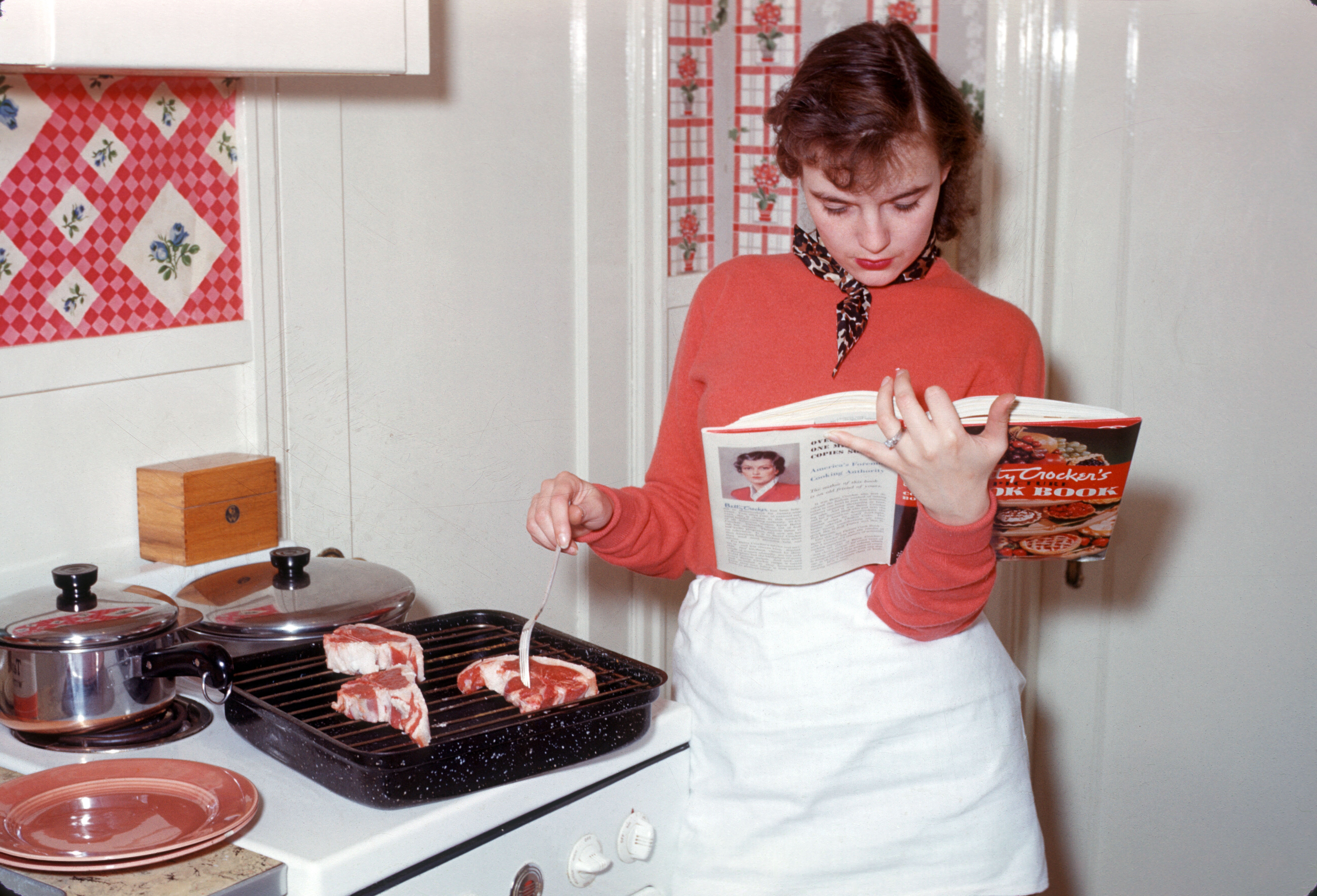 A vintage photo of a woman stirring a pot while reading a cookbook