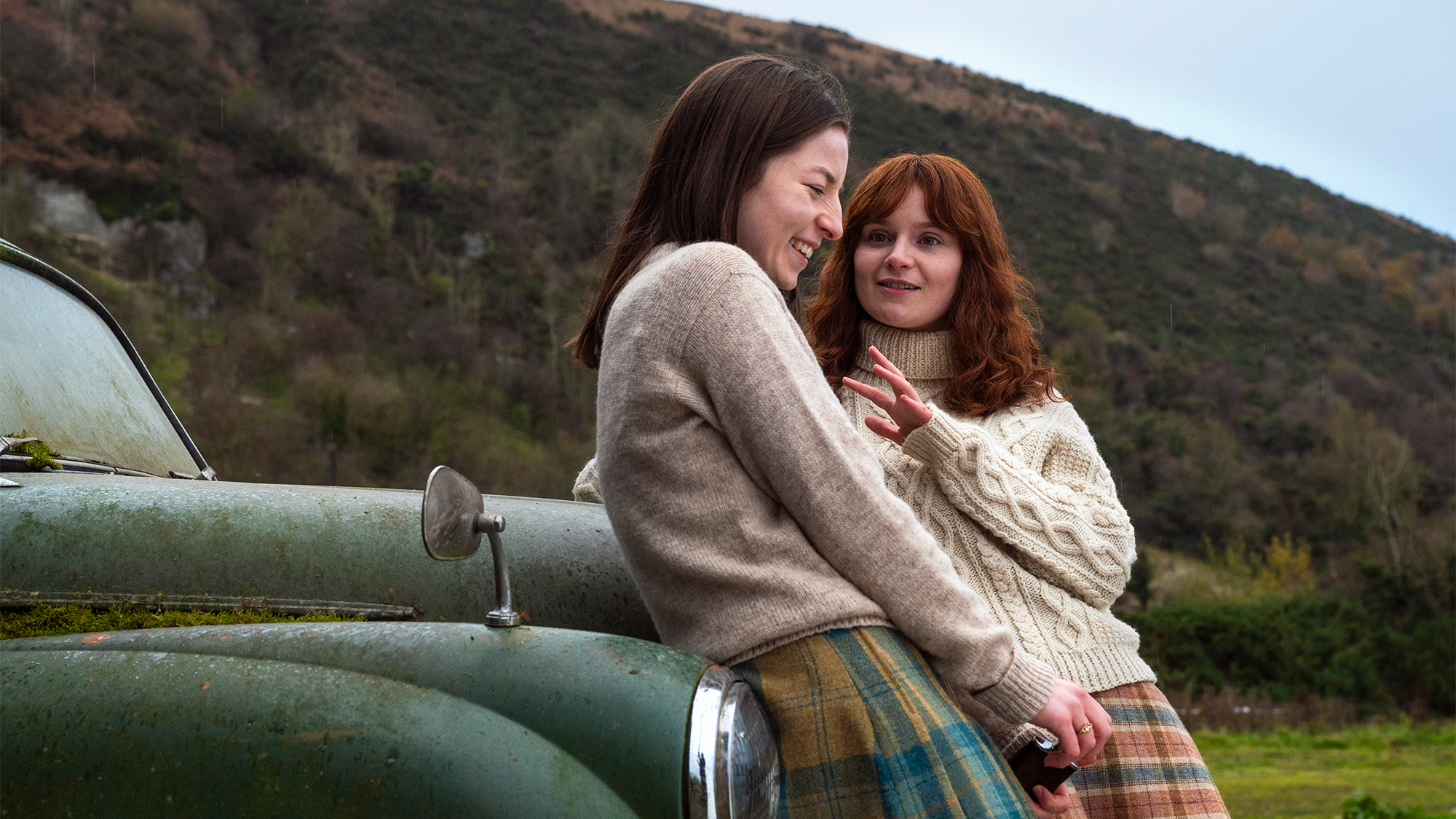 Two women lean on the hood of a car in Ireland and laugh