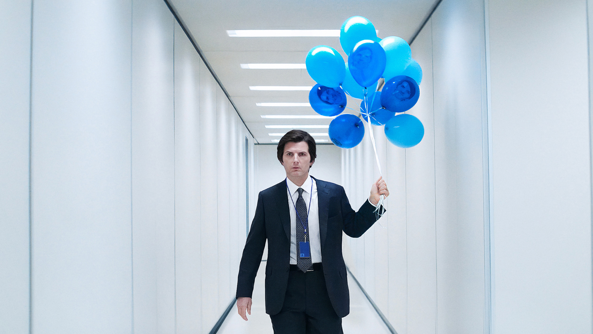 Adam Scott holding blue balloons while walking down a white hallway