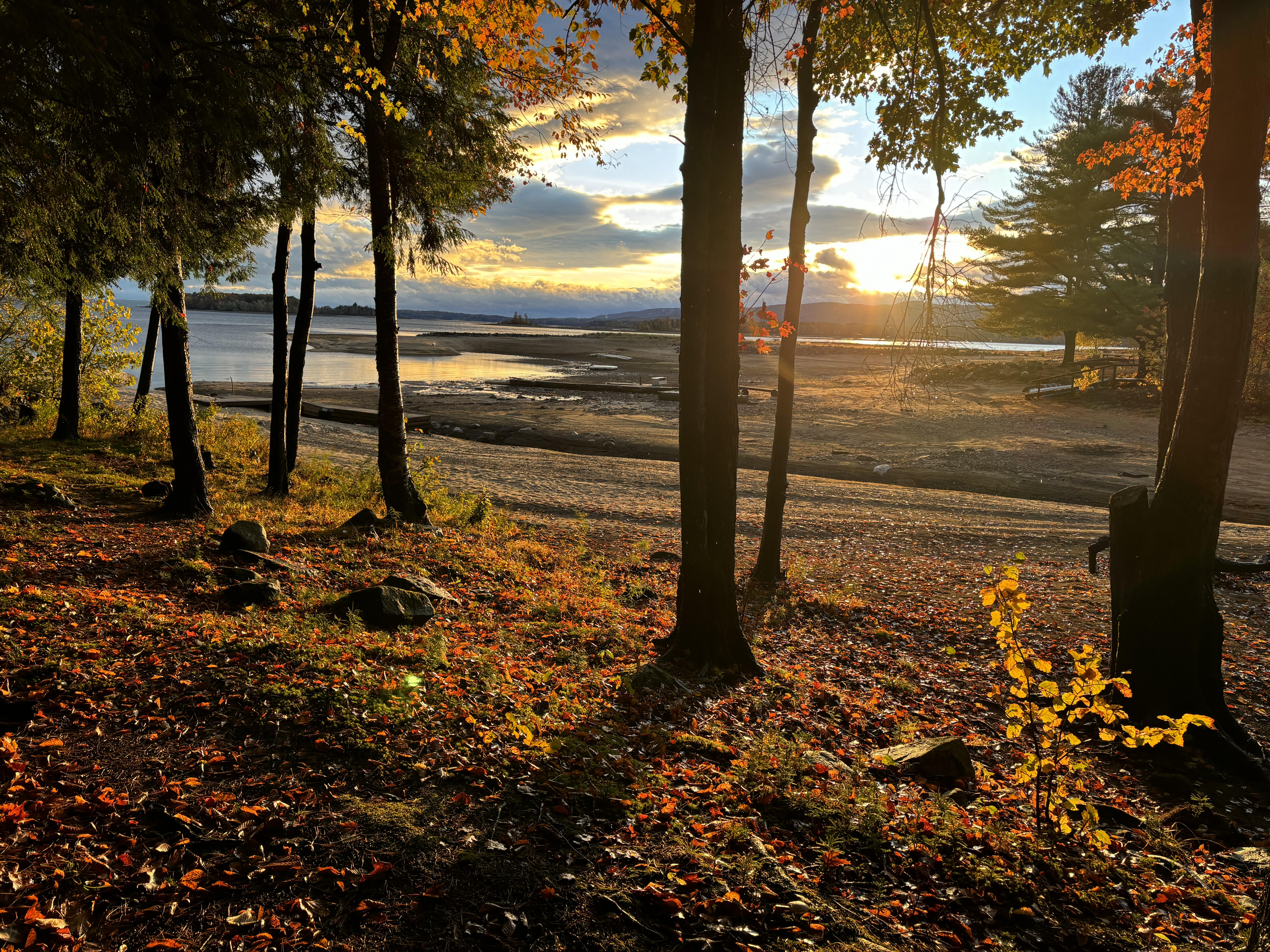 A photograph of trees in red foliage and beyond it the sunset colors on a beach of Great Sacandaga Lake in New York