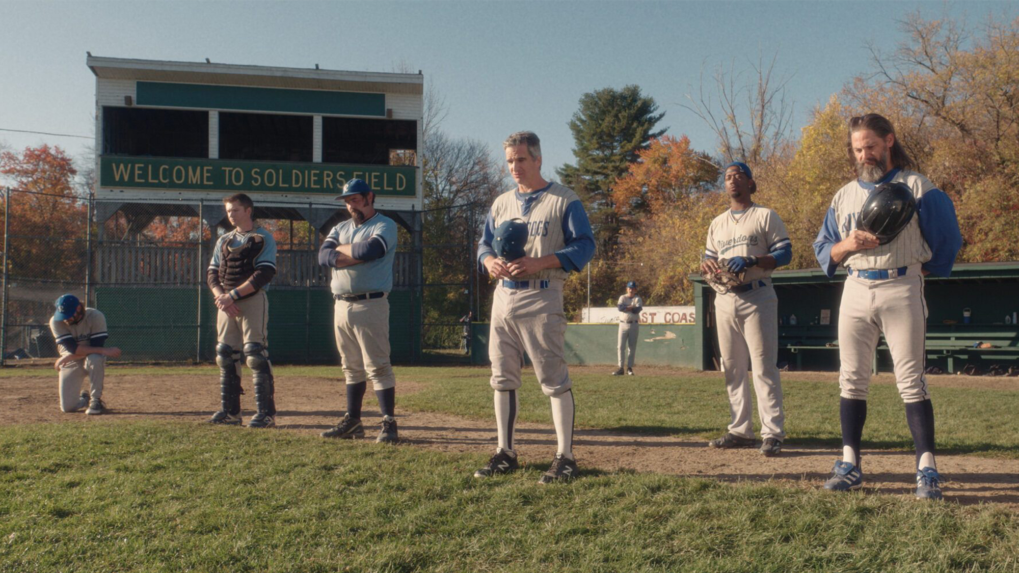 Six men on the baseball team stand on the field with their helmet over their heart