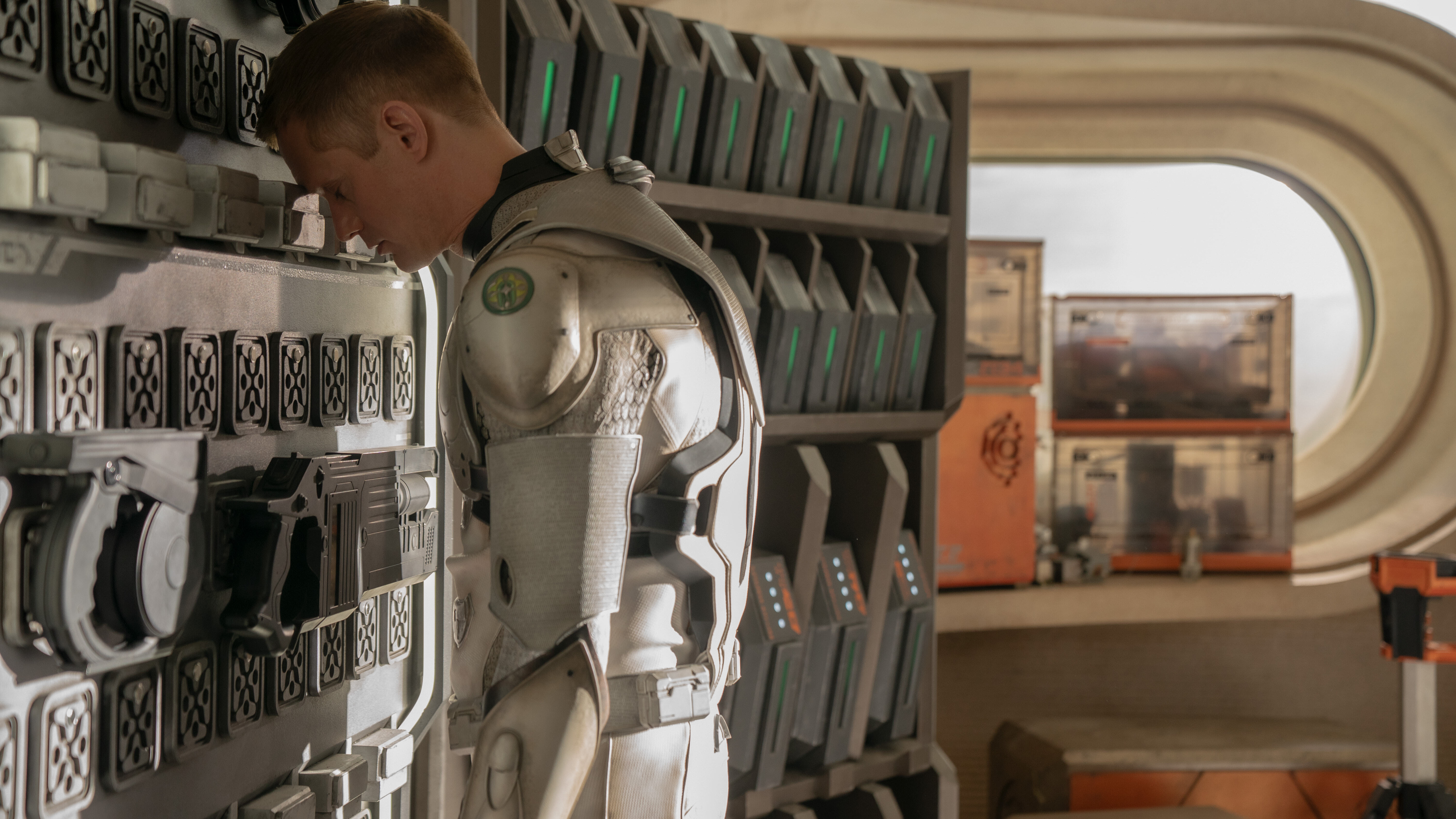 A man in a robotic suit rests his head against a wall of sci-fi tools