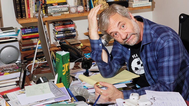 Color photo of Judd Apatow at a cluttered desk with his head in his right hand hand, with bookshelves behind him.