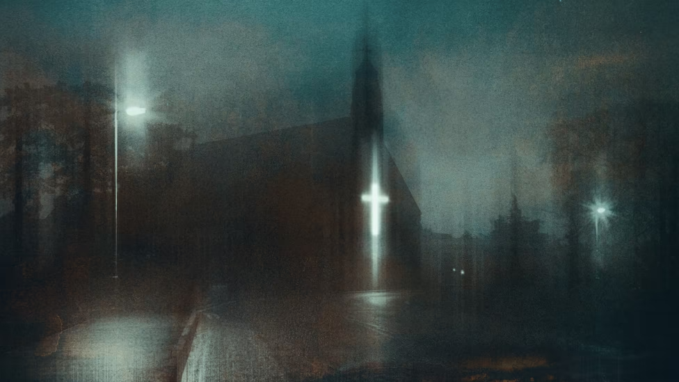 Una fotografía en color de una iglesia con una luz cruzada brillante que se ve desde el otro lado de la calle en una sombría noche azul.