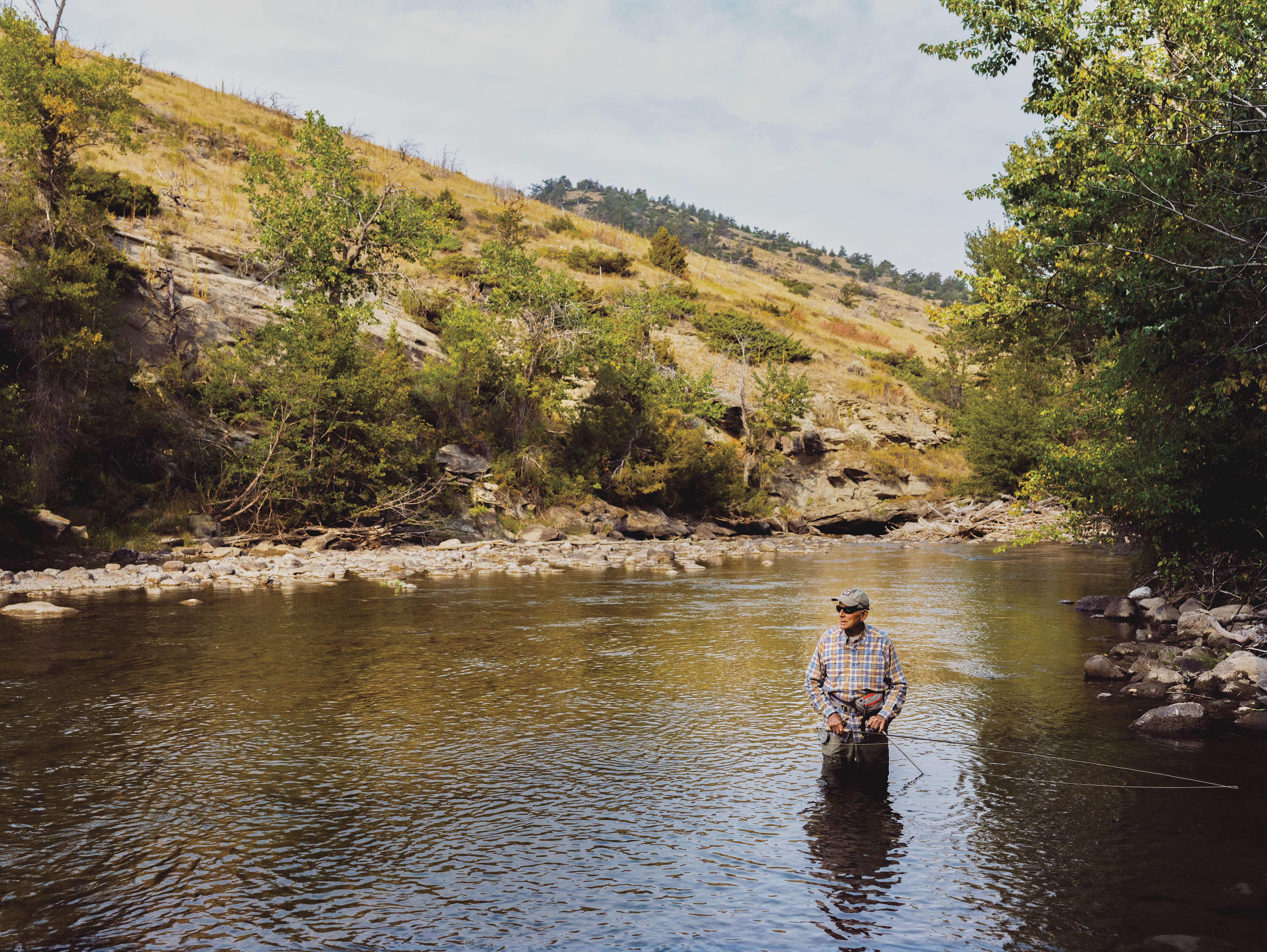 Color photo of a man in waders and a plaid shirt standing knee-deep in a stream holding a fly-fishing rod with hills in the background.