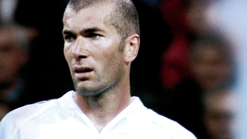 A man with a close-shaven head looking left, in a white soccer jersey, against a blurred background