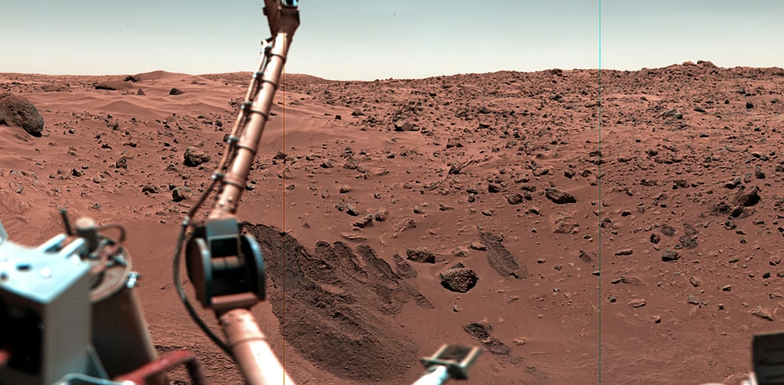 Viking 1’s meteorology instrument stands aloft, on an extended boom, seen against the rocky, sandy surface of Mars.