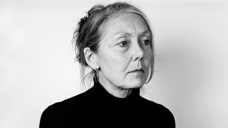 A black-and-white photograph of Nobel Laureate Anne Carson wearing a black turtleneck and hoop earrings, her hair in a messy bun