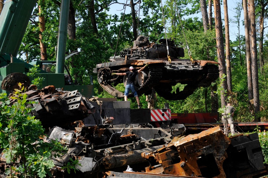 People lift a wrecked tank with a crane, near a pile of wrecked tanks.