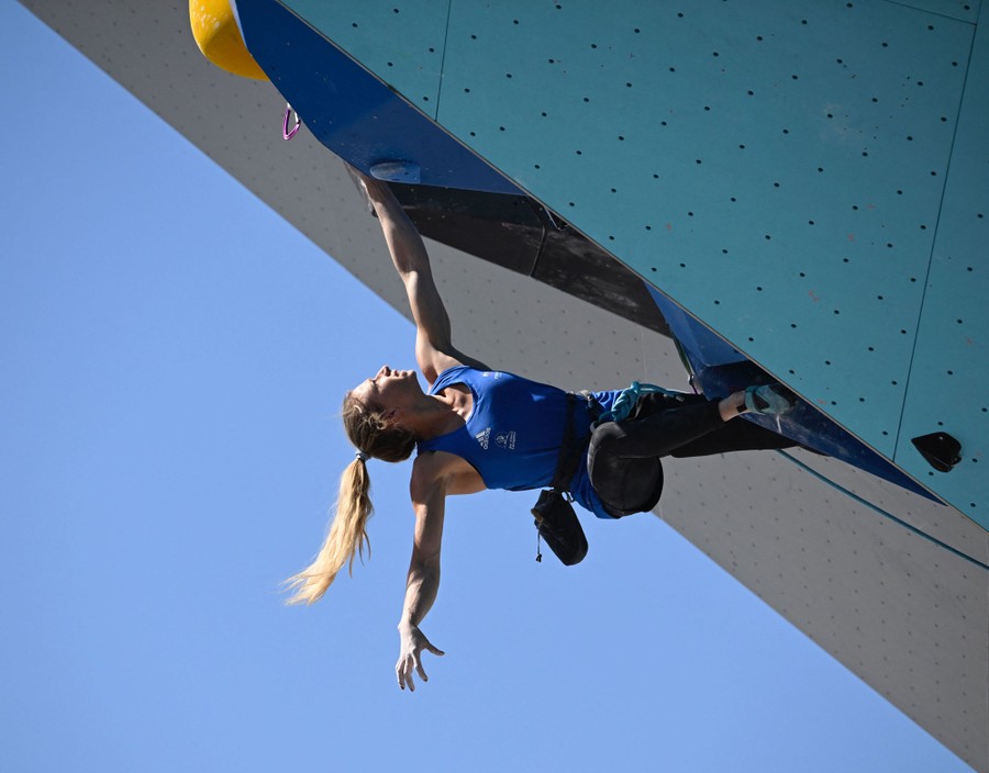 A person competes, hanging nearly upside down, high on a climbing wall.