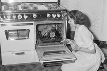 A black-and-white photo of a woman cooking a piece of meat in a gas oven
