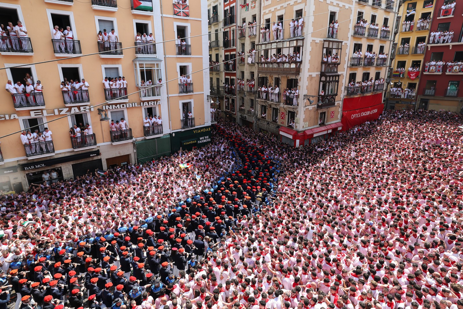 A huge crowd of festivalgoers fills a town square, as a marching band passes through.